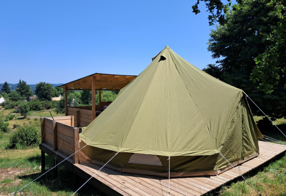 Grande tente cloche installée sur une terrasse en bois avec coin couvert, entourée de verdure et ciel bleu.