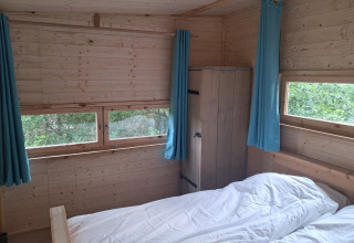 Vista interna di un bungalow in legno con tende azzurre, letto e armadio al camping Junora, Avignone.