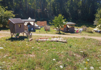 Photo d’un camping de lodge avec cabanes en bois, tentes et sièges extérieurs entourés de verdure et forêt.