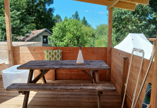 Terrazza in legno con tavolo, panca e cuscino nell’Emperor Bell Tent al Camping Junora, circondato da alberi.