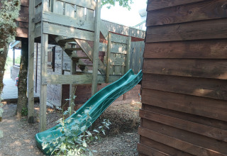 Boomhut met groene glijbaan bij «Eco-Chalet EXTRA» op Camping Junora in Frankrijk, omgeven door bomen.