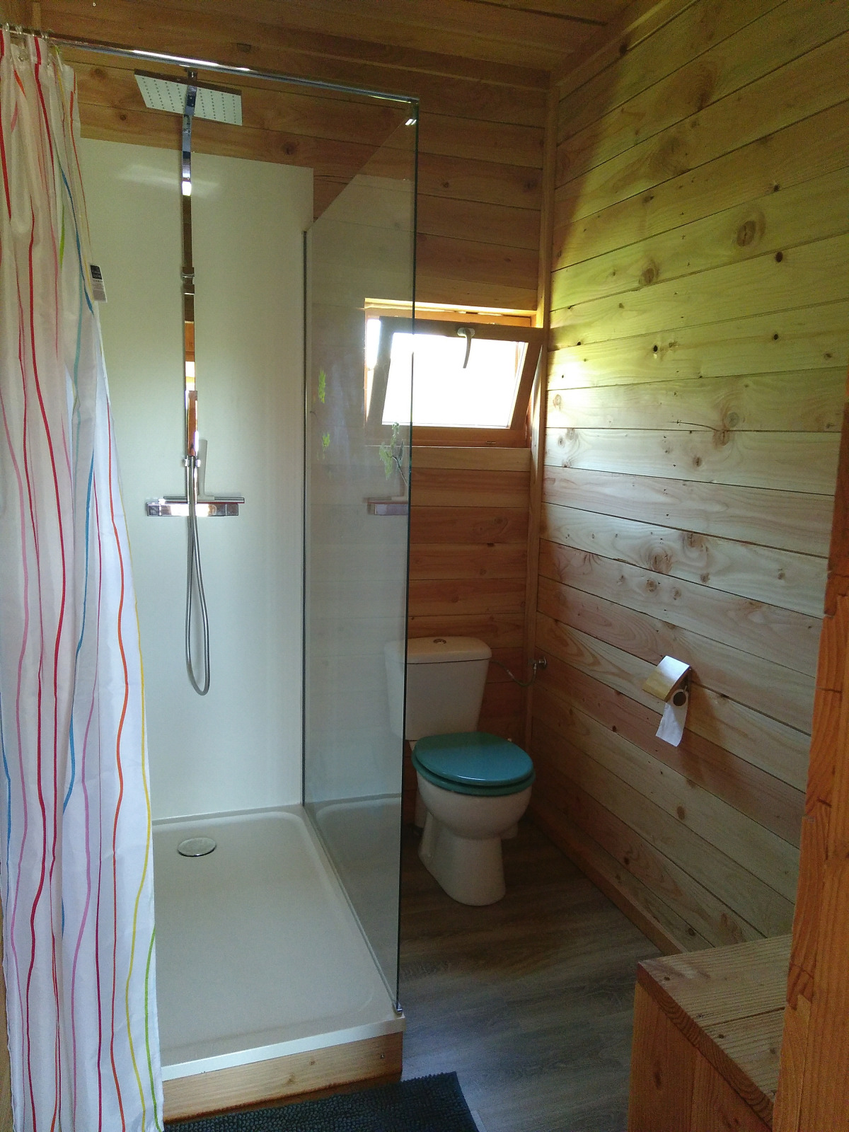 Houten badkamer met douche, toilet met blauwe deksel en een raam in een glamping accommodatie.