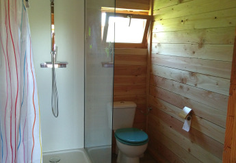 Salle de bain en bois avec douche, toilettes à couvercle bleu et petite fenêtre en hébergement glamping.
