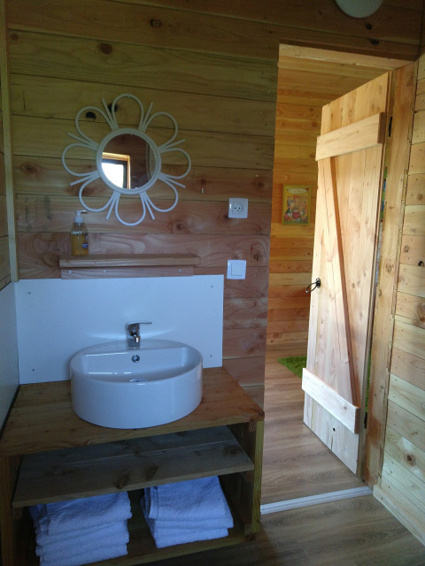 Bagno in legno con specchio rotondo e lavabo nell’Eco-Chalet EXTRA al Campeggio Junora, Francia.