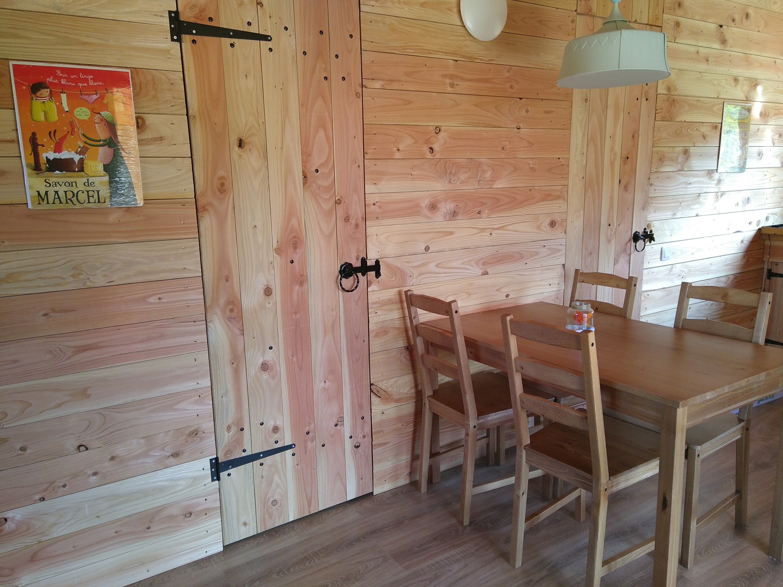 Eetkamer met houten wanden, vier stoelen, tafel, poster en lamp in een luxe zelfvoorzienend eco-chalet.