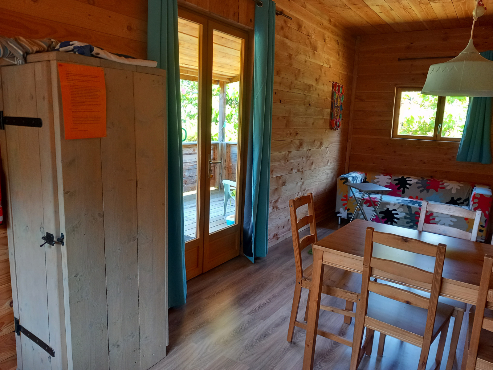 Interieur van een gezellige houten chalet met eettafel, kleurrijke bank en een terras zichtbaar door glazen deuren.