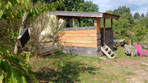 Wooden shelter with chairs and greenery at Bell tent 6M, Campsite Junora, France, on a sunny day.
