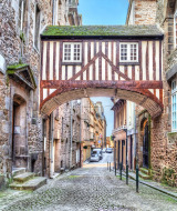 Puente de casa con entramado de madera sobre calle adoquinada rodeada de edificios de piedra en La Chapelle-aux-Filtzméens, Bretaña, Francia.