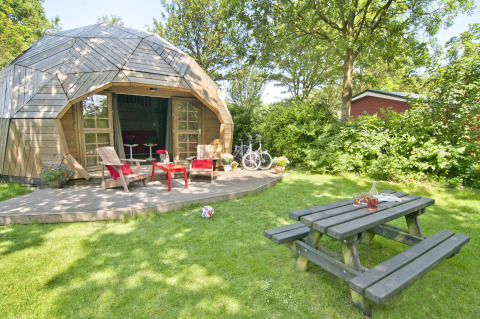 Tiny house igloo di legno con terrazza, sedie da giardino, tavolo da picnic, biciclette e prato verde.