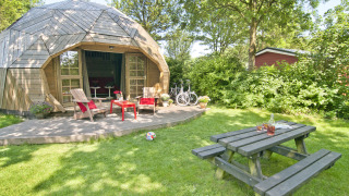 Pequeña casa iglú de madera con terraza, muebles de jardín, bicicleta y mesa de picnic en el césped.