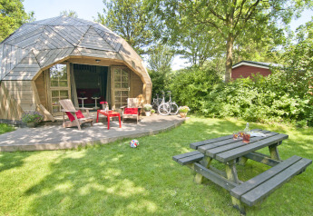 Pequeña casa iglú de madera con terraza, muebles de jardín, bicicleta y mesa de picnic en el césped.