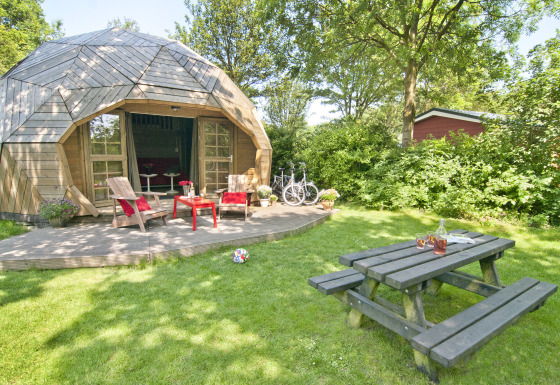 Træiglo tiny house med terrasse, havemøbler, picnicbord, cykler og grøn have på en solrig dag.