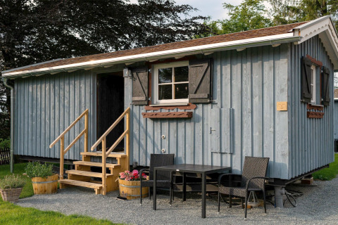 Tiny house bleue en bois avec escalier et coin salon extérieur, prise dans un jardin verdoyant en journée.