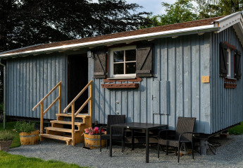 Tiny house bleue en bois avec escalier et coin salon extérieur, prise dans un jardin verdoyant en journée.