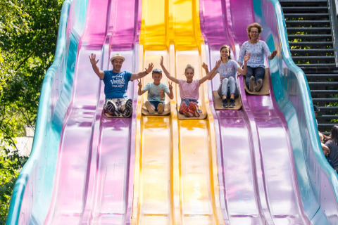 Five people, both children and adults, slide down a large colorful outdoor slide together in the sun.