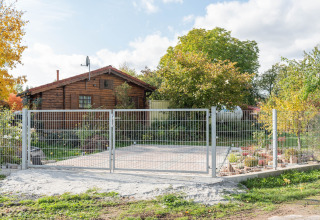 Træhytte i Tyskland med en metalport foran, omgivet af efterårstræer og en have under en blå himmel.