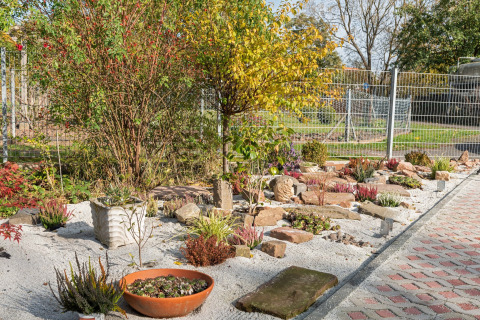 Solrig have i Niederaula, Tyskland, med sand, planter, buske og sten omgivet af metalhegn og sti.