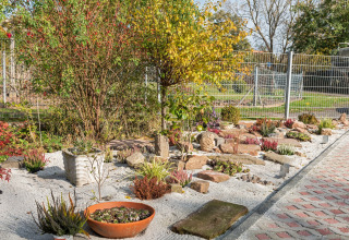 Solrig have i Niederaula, Tyskland, med sand, planter, buske og sten omgivet af metalhegn og sti.