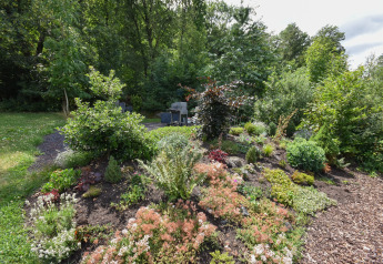 Jardin luxuriant avec arbustes et fleurs variés, entouré d’arbres et d’un coin salon extérieur proche.