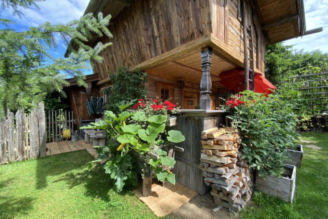 Rustieke Almhütte chalet met houten gevel, bloemen, houtstapel en groene tuin onder een blauwe lucht.