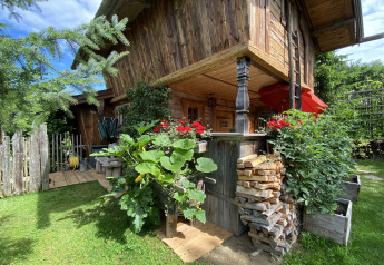 Rustic Almhütte chalet with wooden exterior, flowers, stacked firewood, and lush garden on a sunny day.