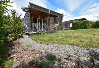 Træhytte med terrasse og liggestole på græsplæne, grussti og blomsterbede under blå himmel i Tyskland.
