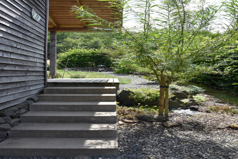 Steps leading to a wooden cabin entrance with gravel path, small tree, and greenery in a peaceful setting.