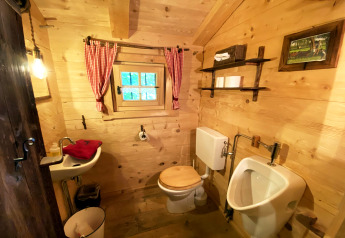 Rustic wooden bathroom at Almhütte chalet featuring toilet, urinal, small sink, and red curtains.