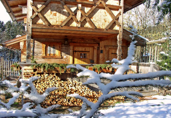 Chalet di legno innevato chiamato 'Almhütte' in Germania, con catasta di legna e rami di pino innevati.