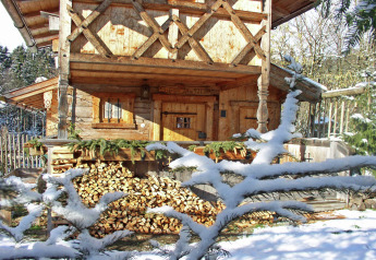 Snow-covered wooden chalet named 'Almhütte' in Germany, with stacked firewood and snowy pine branches.