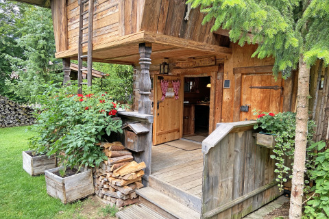 Eingang zu einer rustikalen Berghütte namens „Almhütte“ in Deutschland, umgeben von Pflanzen und Holz.