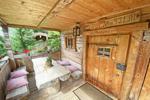 Landelijke houten veranda van een Duitse Almhütte met banken, tafel, kussens en bloemen.