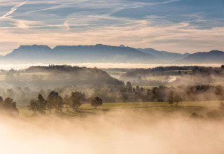 Mistig landschap vanaf chalet Almhütte in Duitsland met bergen, velden en bomen bij dageraad.