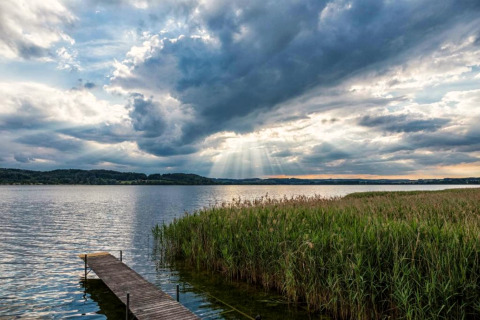Blick auf einen See mit Steg, Schilf und dramatischem Himmel bei Sonnenuntergang nahe Almhütte, Deutschland.