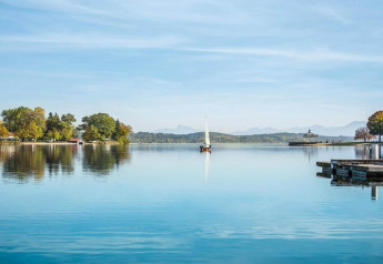 View from Almhütte chalet in Germany showing a sailboat, calm lake, pier, and trees with mountains behind.