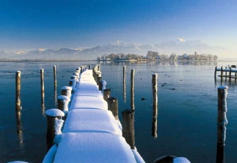 Molo innevato attraversa un lago ghiacciato verso un’isola, montagne sullo sfondo in Germania.