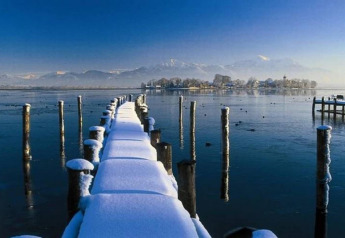 Snow-covered pier stretches over a frozen lake towards an island, alpine mountains in the background, Germany.