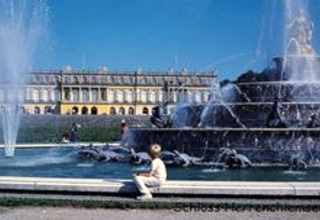 Springbrunnen und Schloss Herrenchiemsee in Deutschland, Besucher sitzen am Wasserrand an einem sonnigen Tag.