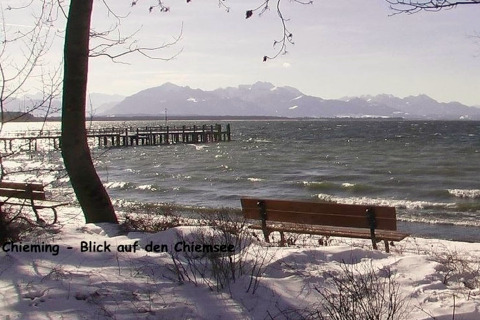Blick von einer schneebedeckten Uferbank am Chiemsee auf das Wasser und die Berge in Deutschland.