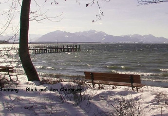Vista del lago Chiemsee da una riva innevata con panche, alberi e montagne sullo sfondo, Germania.