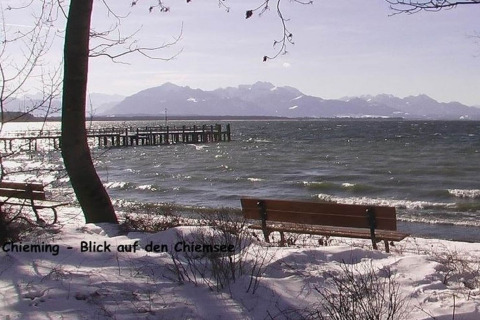 Uitzicht op Chiemsee vanaf een besneeuwde oever met banken, bomen en bergen op de achtergrond, Duitsland.