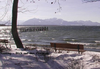Uitzicht op Chiemsee vanaf een besneeuwde oever met banken, bomen en bergen op de achtergrond, Duitsland.