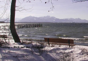 View of Chiemsee lake from a snowy shore with benches, trees, and mountains in the background, Germany.