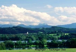 Blick von der Almhütte in Deutschland auf den Waginger-Tachinger See mit grünen Hügeln und Alpen im Hintergrund.
