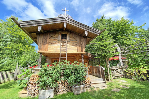 Sfeervol houten Almhütte-chalet met bloemen, brandhout en groen, onder een heldere blauwe lucht.