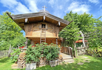 Charming wooden Almhütte chalet with flowers, firewood, and greenery beneath a bright blue sky.