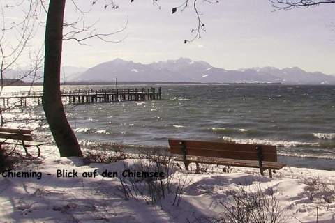 Blick auf den Chiemsee von Chieming aus, verschneiter Strand, Bänke und Berge im Hintergrund.