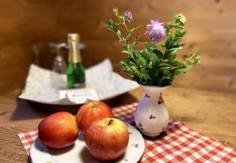 Tavolo rustico di chalet con tre mele rosse, vaso di fiori viola e bottiglia di champagne sullo sfondo.