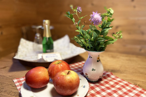 Gezellige chalet-tafel met drie rode appels, vaas met paarse bloem en fles champagne op de achtergrond.