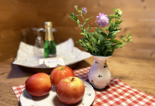 Gezellige chalettafel met drie rode appels, vaas met paarse bloem en fles champagne op de achtergrond.